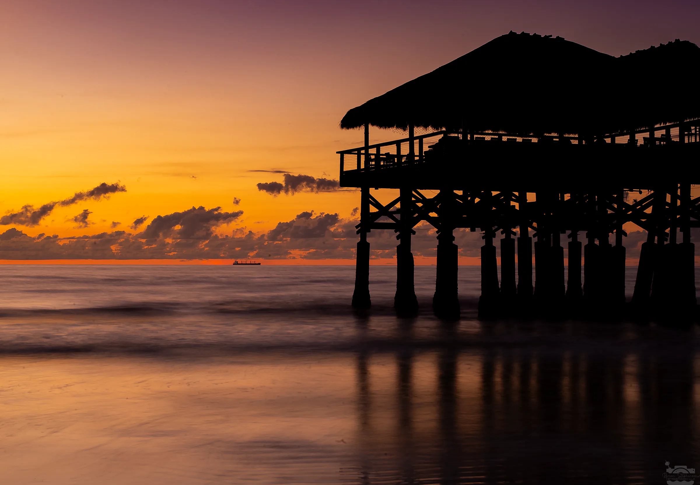 Silhouetted wooden pier with thatched roof at sunset, ocean, and distant ship.