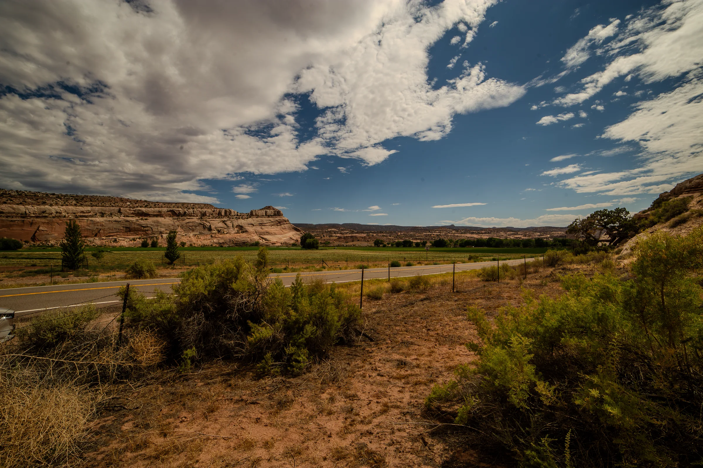 A rural highway curves through a desert valley with sagebrush and distant cliffs.
